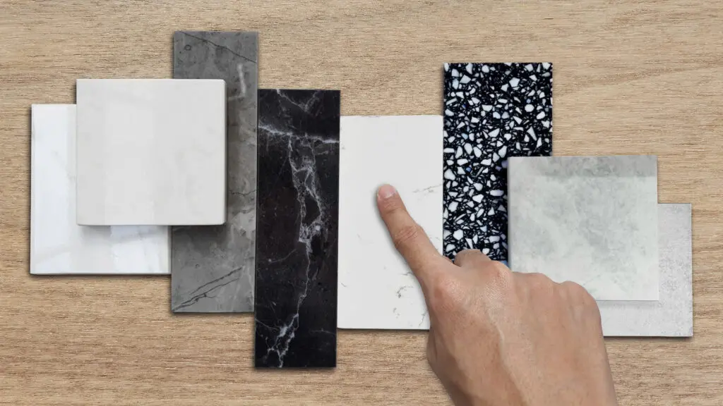 interior designer's hand selects premium stone samples including black marble, grey artificial stone, terrazzo tiles placed with floor tiles. counter top samples on wooden table background.