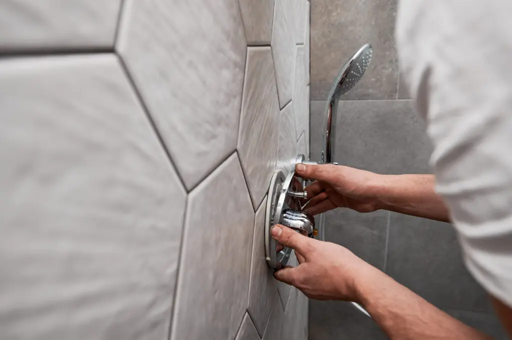 Close up of man standing by the wall with ceramic tile and installing shower faucet with metal handle in apartment. Male plumber working on bathroom renovation at home. Plumbing works concept.